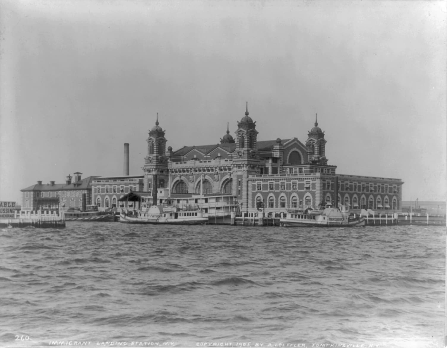 Black-and-white photograph, 1905: the immigrant landing station at Ellis Island seen from the water, ferries moored at the dock